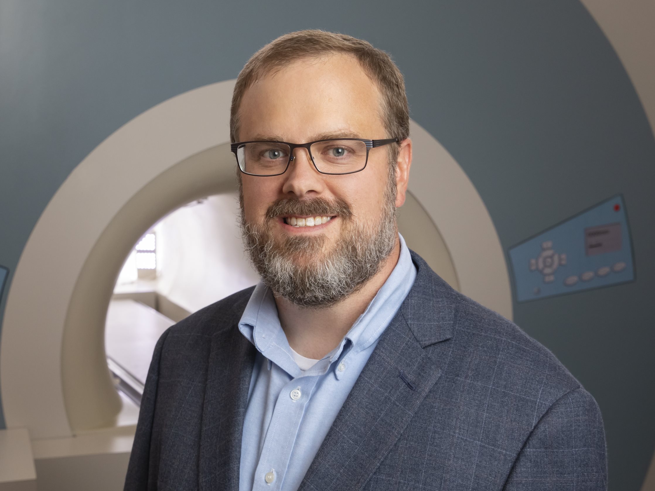 A man smiles at the camera while standing in front of an MRI machine
