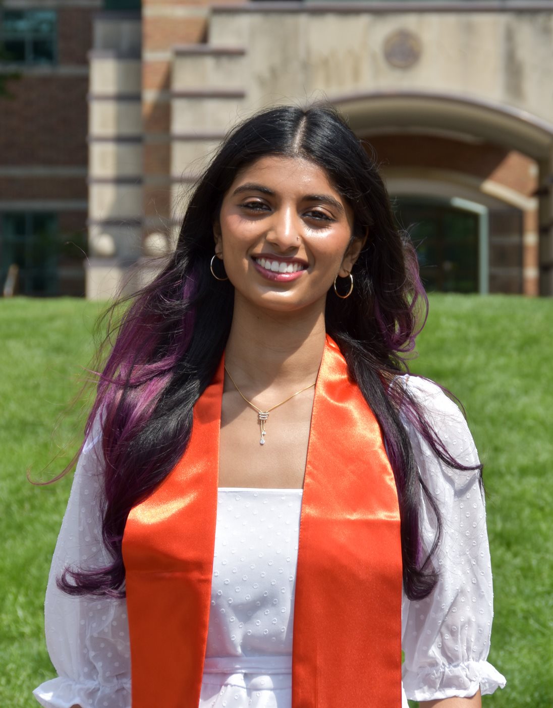 A student smiles at the camera while outdoors wearing an orange graduation sash.