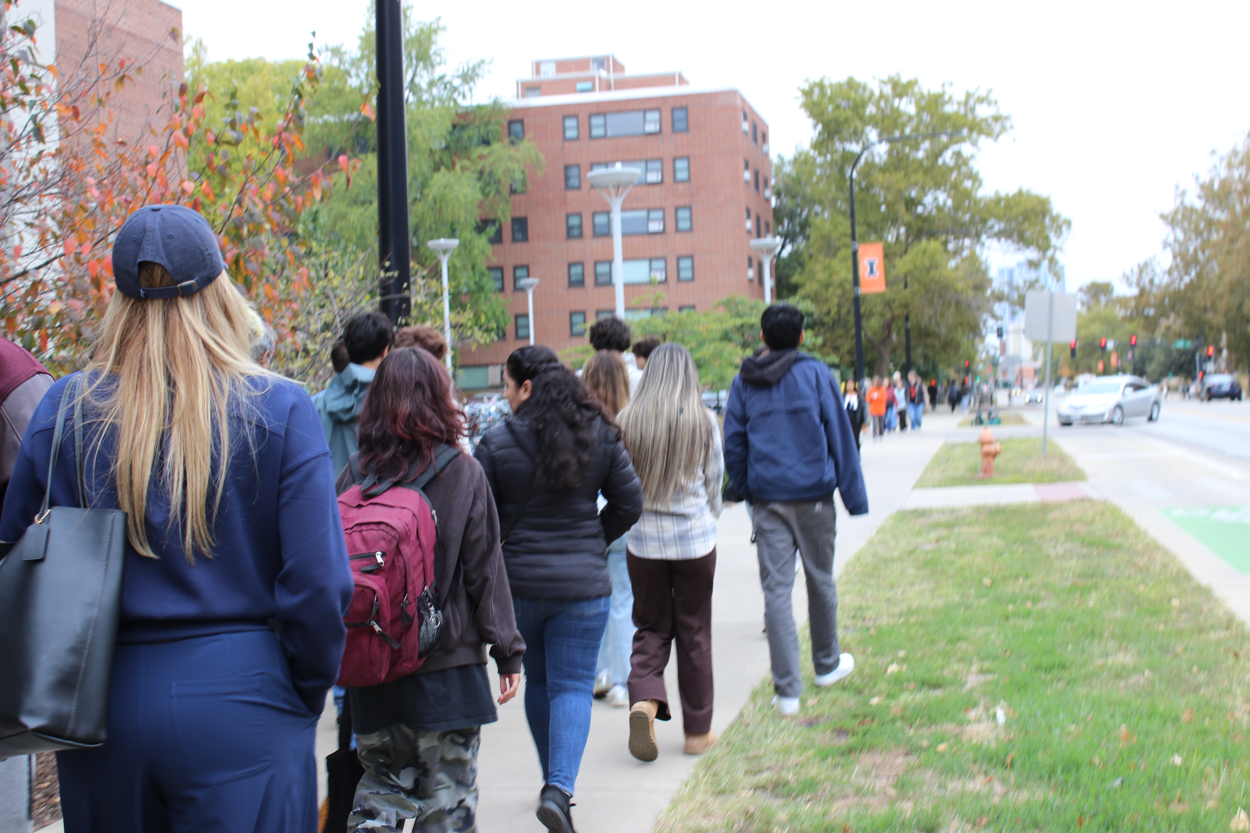 A line of students walk down a sidewalk with a block I banner hung from a lightpole in front of them.
