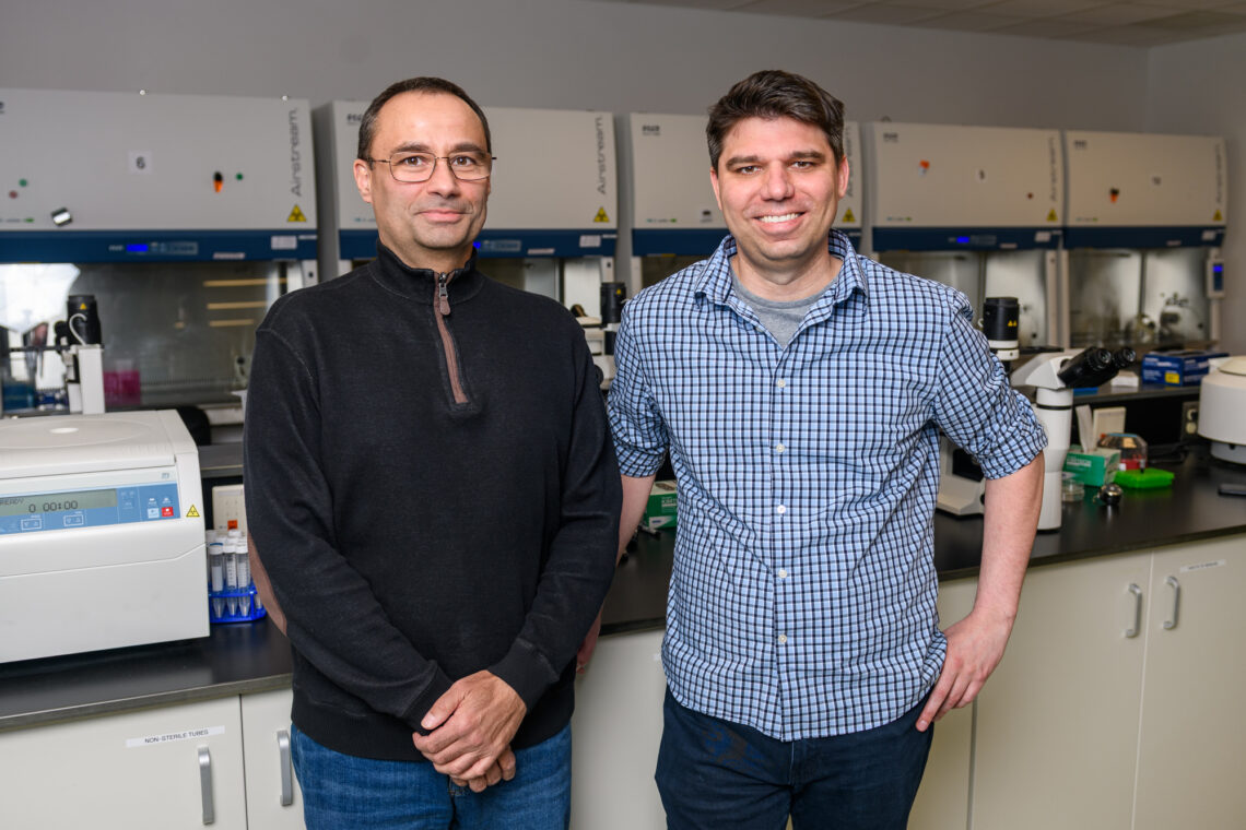 Two men stand side by side in a lab and smile.
