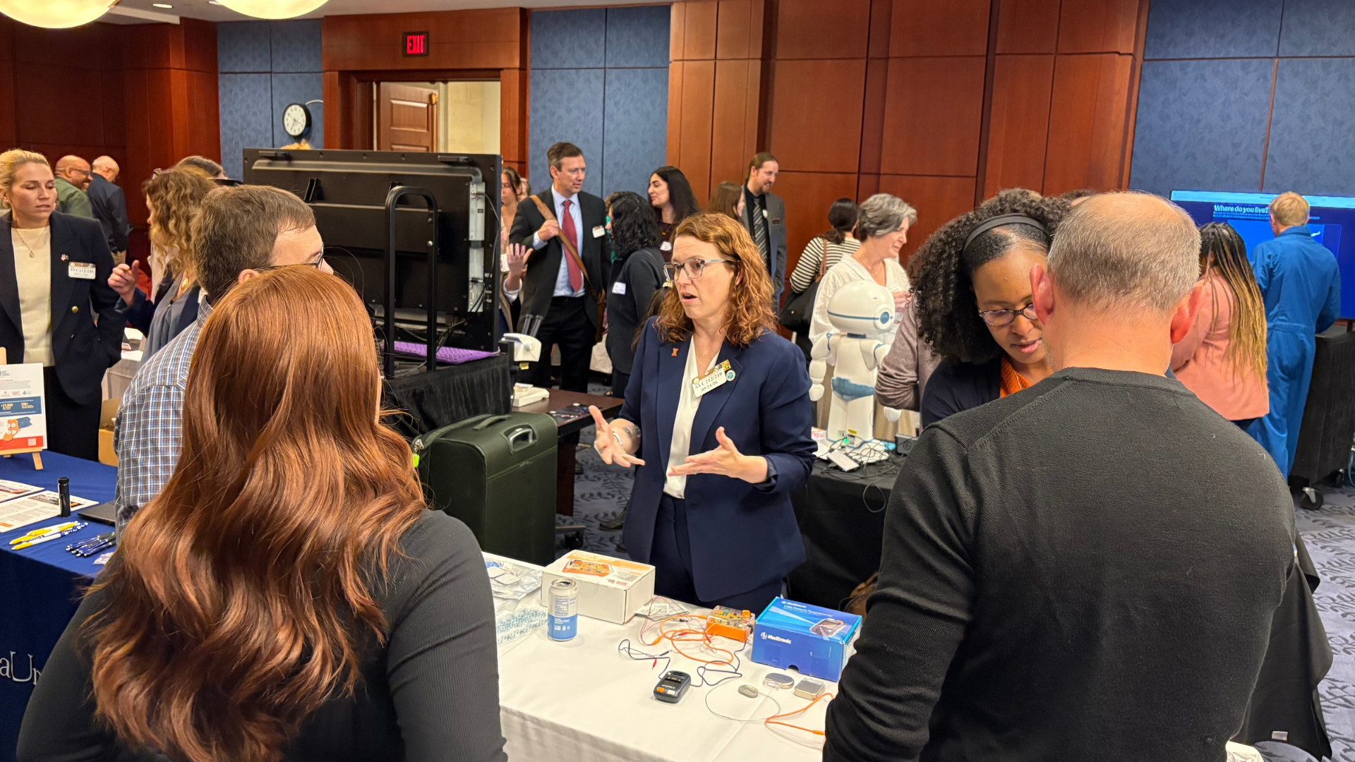 A woman in a blue jacket stands at a table and discusses science to a rapt crowd.